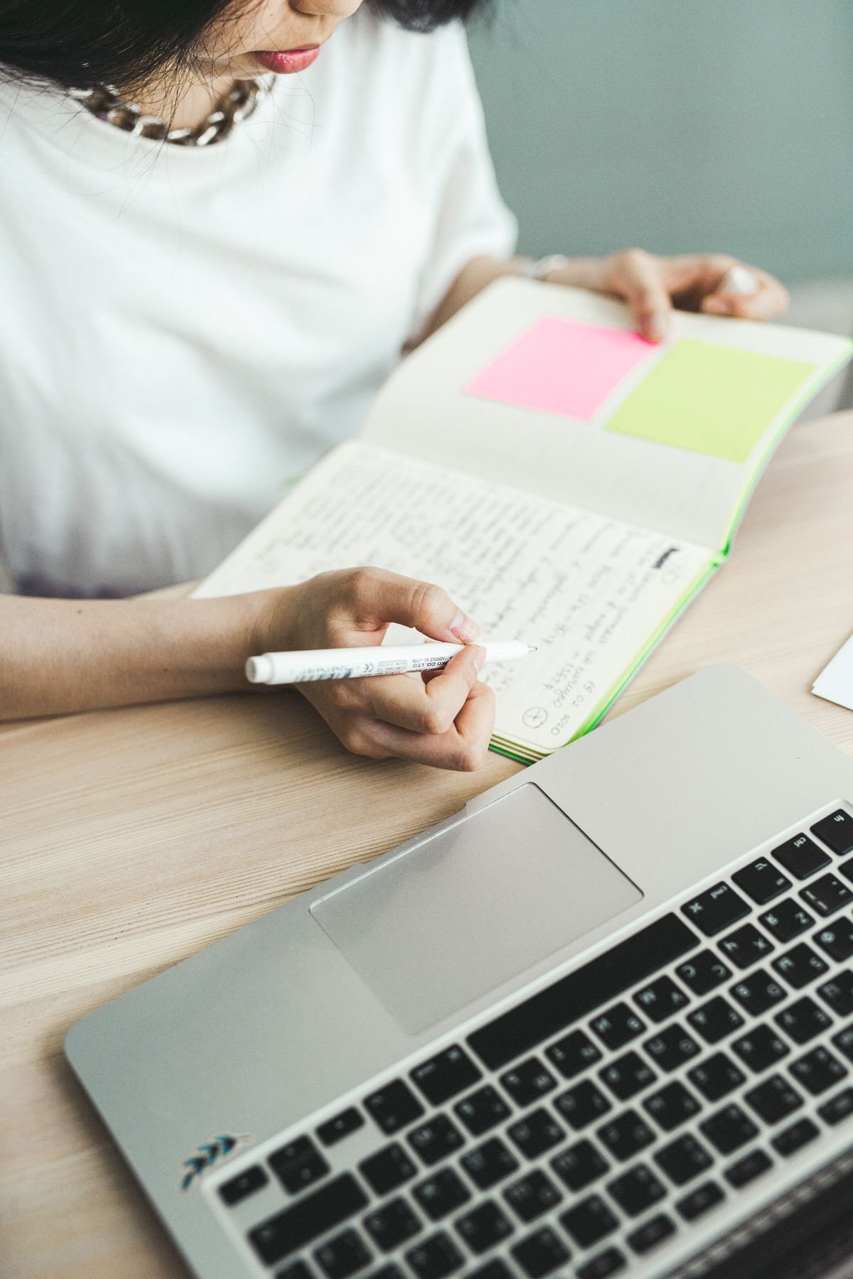 Close-up of a person writing notes in a notebook beside a laptop