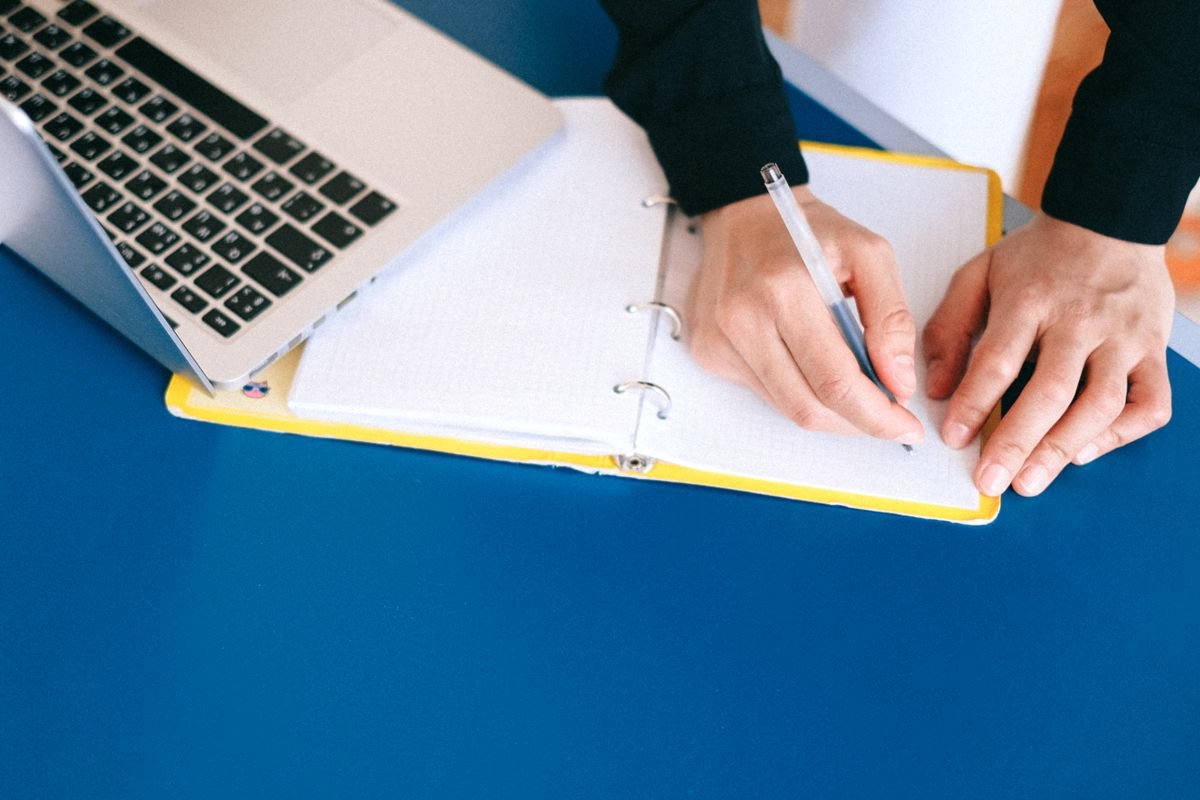 Hands writing in a notebook with a laptop nearby
