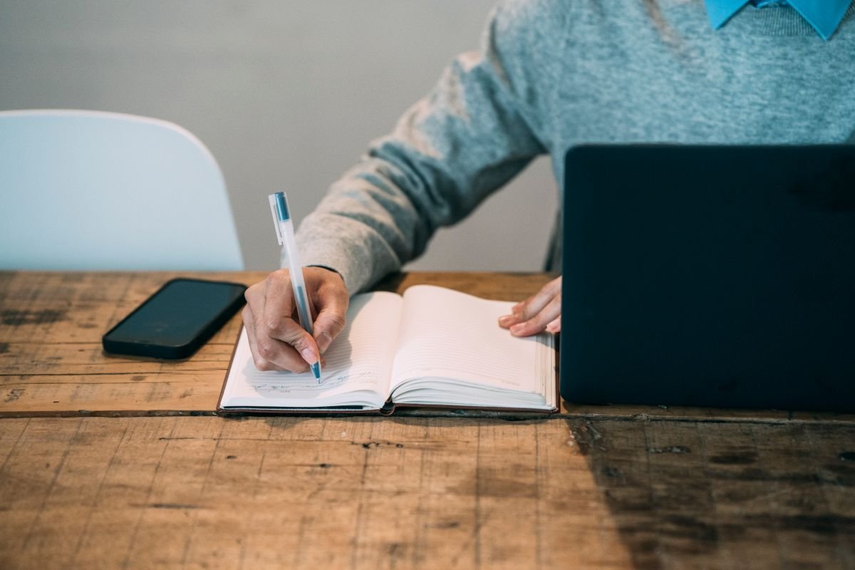 Person writing notes in a notebook beside a laptop